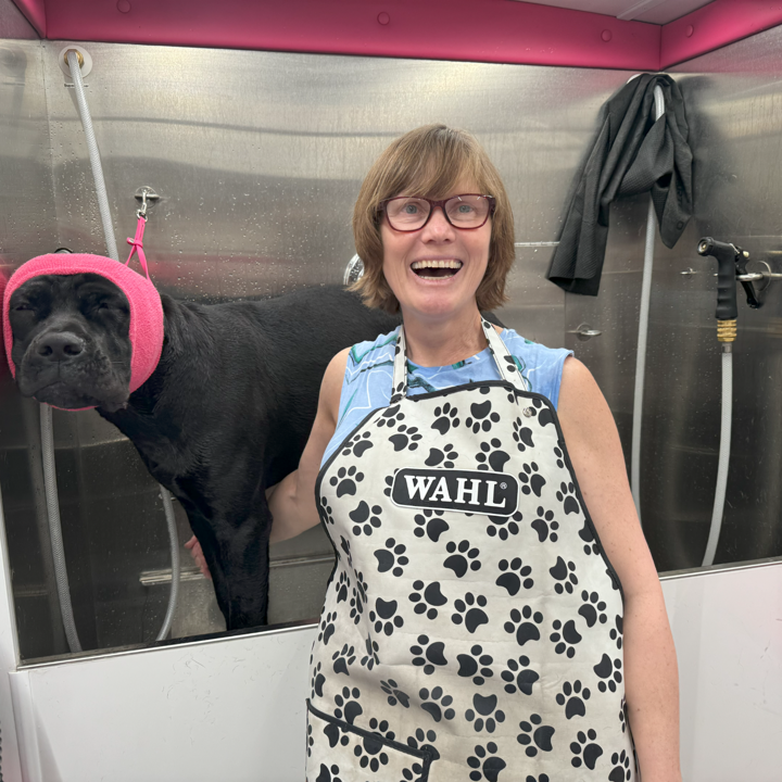 Helen, Lead Groomer and Co-founder of Bow Tie Mobile Pet Grooming, laughing joyfully while bathing a black Cane Corso inside the state-of-the-art mobile grooming van during an at-home grooming appointment in Shaddock Creek Estates, Frisco, TX. The dog wears bright pink ear protection (happy hoodie) to dampen the noise from blow-drying, showcasing Helen's attention to pet comfort and safety. The stainless steel bathing station, Super Sudser bathing system (to create a massaging, thorough clean), and commercial-grade variable-speed blow dryer reflect the fully-equipped mobile salon that brings stress-free, one-on-one grooming directly to homes throughout Frisco, Plano, McKinney, Prosper, and The Colony. With over 10 years of experience, Helen specializes in fear-free handling for anxious dogs, nervous rescues, and senior pets.